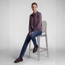 Load image into Gallery viewer, A woman sits on a clear acrylic bar stool wearing a dark purple and black patterned button-down shirt, blue jeans, and brown leather loafers against a light gray studio background.
