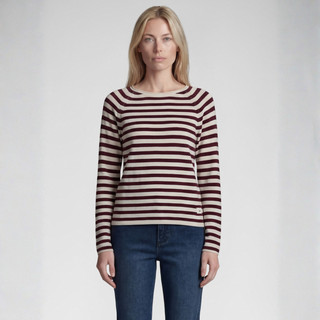 A woman modeling a long-sleeve knit top with horizontal burgundy and cream stripes, paired with dark blue jeans, against a light gray studio background.