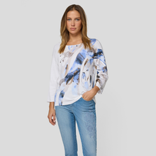 Load image into Gallery viewer, Woman wearing a white blouse with blue abstract patterns and blue jeans on a light gray background
