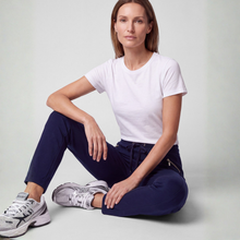 Load image into Gallery viewer, A woman sits cross-legged on a light gray studio floor wearing a plain white short-sleeve T-shirt, navy jogger-style pants with a small gold zipper detail on the pocket, and white New Balance sneakers.
