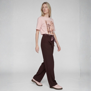 A woman modeling a light pink graphic t-shirt and dark brown wide-leg trousers with a matching patterned waist tie, paired with light pink loafers, against a light gray studio background.