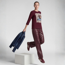 Load image into Gallery viewer, A model jumps over a white block in a studio setting, wearing a burgundy long-sleeve graphic t-shirt, matching burgundy faux-leather pants, burgundy sneakers, and holding a blue denim jacket.
