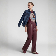 Load image into Gallery viewer, A model is shown from the waist down wearing a dark blue denim jacket, a burgundy graphic t-shirt, and matching burgundy faux-leather wide-leg pants with burgundy sneakers, set against a plain light gray studio background.

