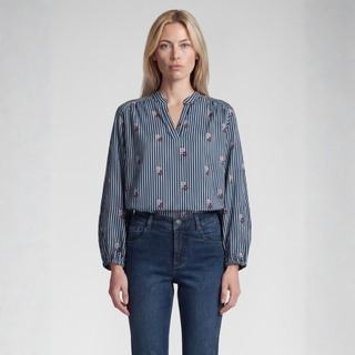 A woman modeling a long-sleeve button-up blouse with vertical navy and white stripes and small red floral accents, paired with dark blue jeans, against a light gray studio background.