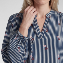 Load image into Gallery viewer, A close-up of a woman wearing a navy and white vertically striped blouse with small floral prints and a tie detail at the collar.
