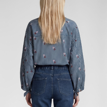 Load image into Gallery viewer, A woman viewed from the back wearing a blue and white striped long-sleeve blouse with small floral embroidery and dark blue jeans, against a light gray studio background.
