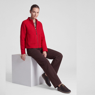 A woman models a bright red zip-up jacket with front patch pockets, paired with dark brown trousers and dark brown sneakers, sitting on a white cube against a light gray studio background.