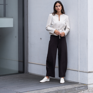 Woman in a white blouse and black pants standing against a gray wall.