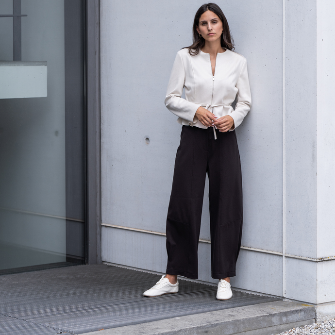 Woman in a white blouse and black pants standing against a gray wall.