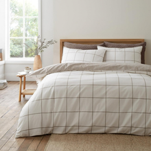 Load image into Gallery viewer, A neatly made bed in a bright bedroom featuring a cream duvet cover with a brown grid pattern, matching striped pillow shams, and two dark brown patterned pillows. The room has light wood flooring, a wooden headboard, and a small side table by a window.
