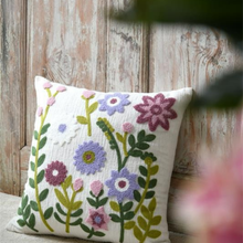 Load image into Gallery viewer, A square white throw pillow with colorful embroidered wildflowers and green leaves, resting against a rustic weathered wood panel background.
