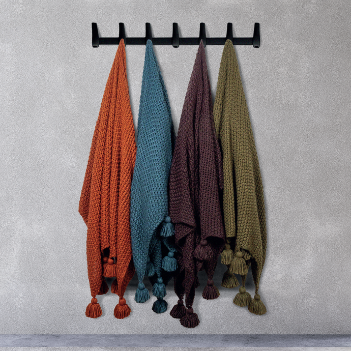 Four Scatterbox throws hanging on a grey wall from black hooks, featuring orange, blue, aubergine and green