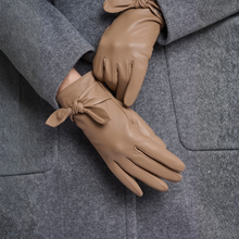 Load image into Gallery viewer, A pair of tan leather gloves with wrist bows, worn on hands resting on a gray wool coat.

