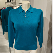 Load image into Gallery viewer, A teal blue long-sleeve knit polo sweater displayed on a white mannequin torso in a clothing store. The sweater features a classic collar, three decorative buttons on the placket, and a geometric embroidered or stitched pattern on the chest.
