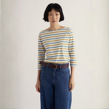 Load image into Gallery viewer, A woman with short black hair stands against a plain off-white background wearing a three-quarter sleeve striped top with yellow, blue, and white horizontal stripes, paired with high-waisted blue jeans and a brown belt.
