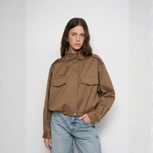 Load image into Gallery viewer, A woman modeling a brown utility jacket with a high collar, drawstrings, and flap chest pockets, paired with light blue jeans, against a plain light gray studio background.
