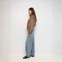 Load image into Gallery viewer, A woman stands in profile wearing a brown long-sleeve button-up shirt, wide-leg light blue denim jeans, and black shoes against a plain white studio background.
