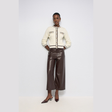 Load image into Gallery viewer, A model stands against a light gray studio background wearing a cream cropped jacket with dark brown trim and matching dark brown wide-leg faux-leather pants, paired with dark brown pointed slingback heels.
