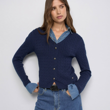 Load image into Gallery viewer, A woman modeling a navy blue cable-knit cardigan with gold buttons over a light blue denim shirt, paired with blue jeans, against a plain light gray background.
