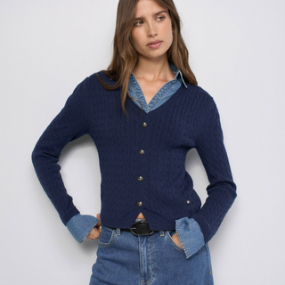 A woman modeling a navy blue cable-knit cardigan with gold buttons over a light blue denim shirt, paired with blue jeans, against a plain light gray background.