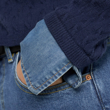 Load image into Gallery viewer, Close-up of a person wearing a navy blue cable-knit sweater layered over a light blue denim shirt or jeans, with the sleeve folded over to show the knit texture and denim fabric.
