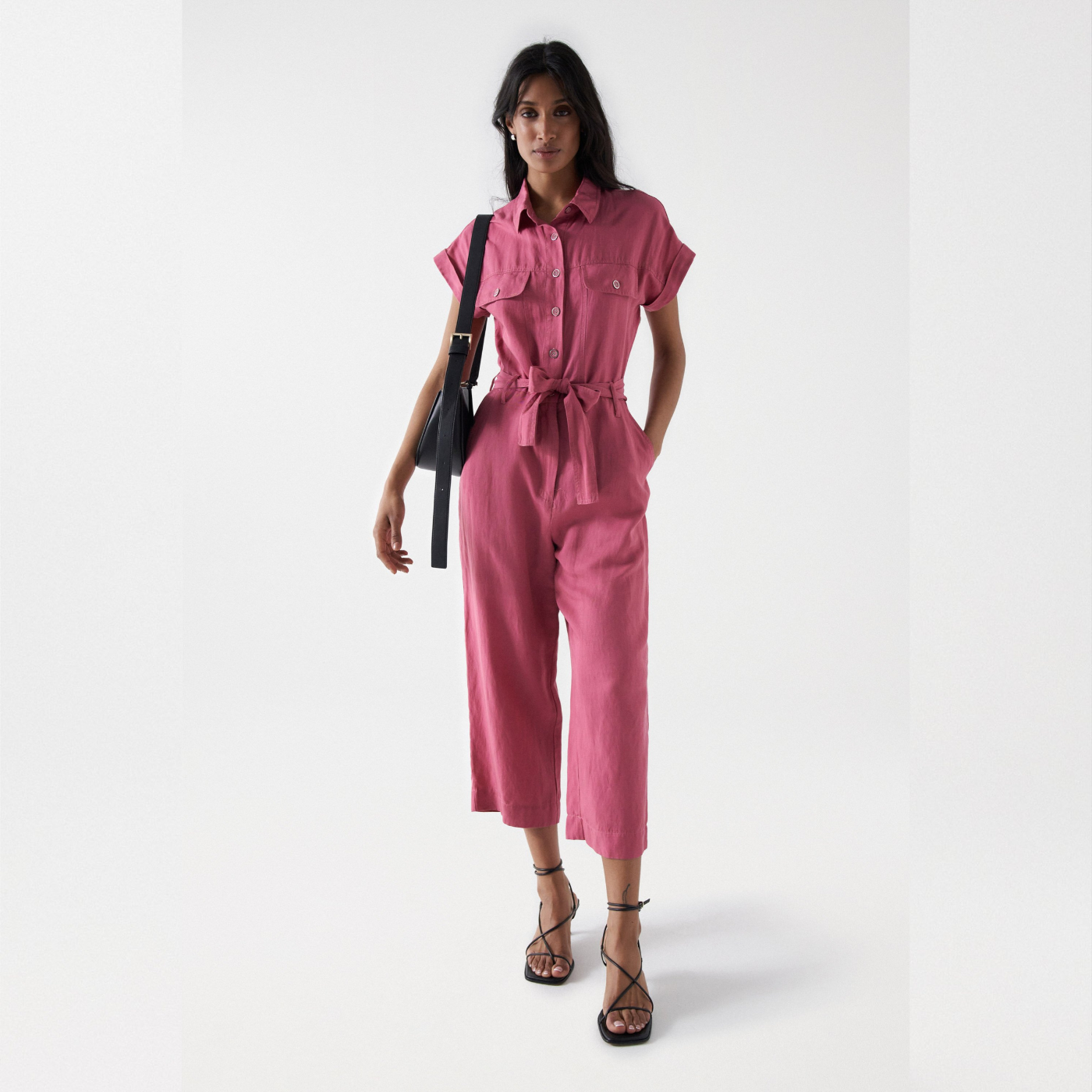 Dark hair model standing. Wearing Salsa jumpsuit in old pink color. Jumpsuit has short sleeves, buttoned top and belt around waist. 