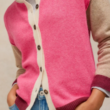 Load image into Gallery viewer, A close-up of a person wearing a pink knit cardigan with cream ribbed trim and dark buttons, layered over a beige long-sleeve top, paired with blue jeans.
