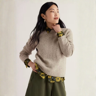 A woman wearing a beige knit sweater over a yellow and green floral collared shirt and olive green pants, posing against a plain off-white background.