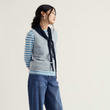 Load image into Gallery viewer, A woman wearing a light gray ribbed knit sweater vest over a light blue and white striped long-sleeve shirt, paired with a dark navy scarf and blue jeans, set against a plain off-white studio background.
