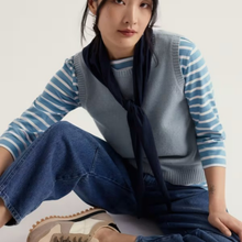Load image into Gallery viewer, A model wearing a light blue knit sweater vest over a blue and white striped long-sleeve shirt, paired with a dark navy neck scarf, blue jeans, and beige sneakers, set against a plain off-white background.
