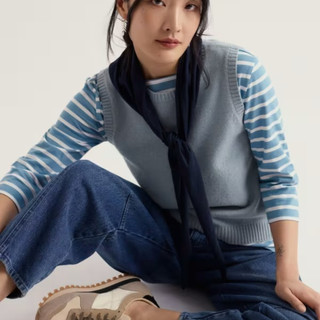 A model wearing a light blue knit sweater vest over a blue and white striped long-sleeve shirt, paired with a dark navy neck scarf, blue jeans, and beige sneakers, set against a plain off-white background.
