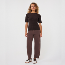Load image into Gallery viewer, A woman stands against a plain light gray studio background wearing a short-sleeve dark brown knit top with subtle speckled texture, loose brown pants, and black-and-white sneakers.
