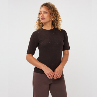 A woman with curly blonde hair wearing a short-sleeve dark brown knit top with subtle speckled details and matching brown pants, standing against a light gray studio background.
