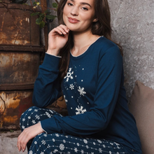 Load image into Gallery viewer, A woman wearing a navy blue long-sleeve pajama top and matching pants with white snowflake patterns, sitting against a rustic brown metal and gray concrete wall.

