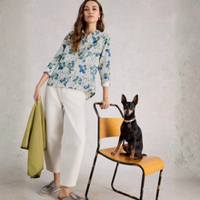 Load image into Gallery viewer, Woman in a floral blouse and white pants holding a green jacket, standing next to a dog on a chair against a gray background.
