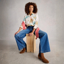 Load image into Gallery viewer, Woman sitting on a wooden box wearing a floral shirt, blue jeans, and brown boots against a plain background
