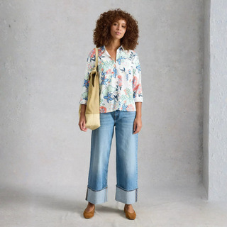 Woman wearing a floral blouse and blue jeans, holding a beige bag against a gray background