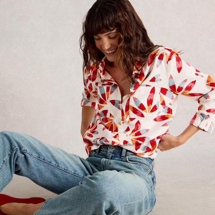 Woman wearing a colorful patterned shirt and jeans sitting on the floor against a plain background