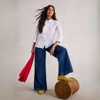 Woman in a white polka dot shirt and blue pants holding a red jacket, standing on a wooden stool against a plain background.