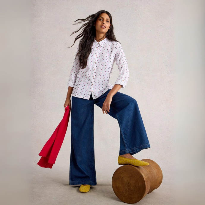 Woman in a white polka dot shirt and blue pants holding a red jacket, standing on a wooden stool against a plain background.
