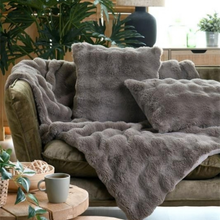 Load image into Gallery viewer, A cozy living room scene with a plush taupe-gray faux fur throw blanket and matching pillows draped over an olive green sofa, with a mug on a wooden side table in the foreground.
