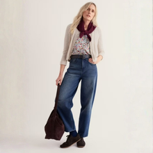 Load image into Gallery viewer, A woman models a stylish outfit featuring high-waisted, wide-leg blue jeans with a brown belt, a cream cardigan, a floral top, a burgundy scarf, and dark brown shoes, while holding a dark brown tote bag against a plain off-white studio background.
