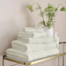 Load image into Gallery viewer, Stack of almond towels on a gold shelf with a vase of greenery in the background.
