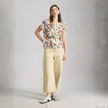 Load image into Gallery viewer, A woman stands against a textured gray studio wall wearing a short-sleeve V-neck blouse with a multicolor abstract print on a light cream background, paired with wide-leg cropped beige pants and black-and-white sandals.
