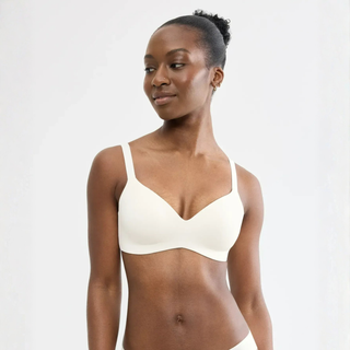 A woman wearing a solid off-white/cream wireless bralette against a plain light gray studio background.