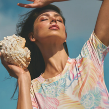 Load image into Gallery viewer, Woman holding a shell with a colorful shirt against a clear blue sky
