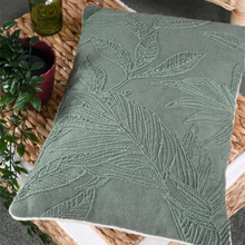 Load image into Gallery viewer, A square sage green throw pillow with an embroidered leaf pattern, styled on a woven wicker tray with a small potted plant and a glass bottle.
