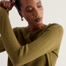 Load image into Gallery viewer, A close-up of a woman wearing a ribbed knit sweater in a muted olive green color, with her hand adjusting her hair against a plain light background.
