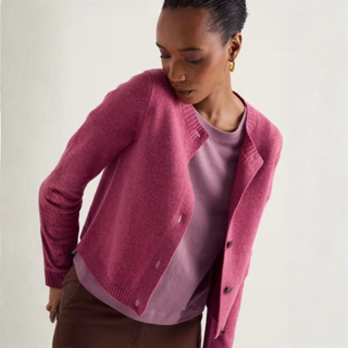 A woman wearing a pink knit cardigan over a mauve top and brown pants, photographed against a plain light background.