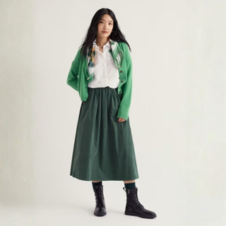 A model stands against a plain off-white studio background wearing a green cardigan, white button-up shirt, dark green midi skirt, and black lace-up ankle boots.